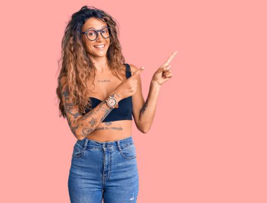 Gündelik giysiler ve gözlük takan, gülümseyen ve kameraya bakan, iki eli ve parmakları yanda olan Latin kökenli genç bir kadın.. 