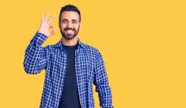 Gündelik kıyafetler giyen genç İspanyol adam gülümsüyor. El ve parmaklarıyla 