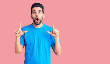 Sakallı, sıradan tişörtlü genç ve yakışıklı bir adam şaşırmış ve şaşırmış bir şekilde yukarı bakıyor ve parmaklarıyla işaret ediyor ve kollarını kaldırıyor.. 