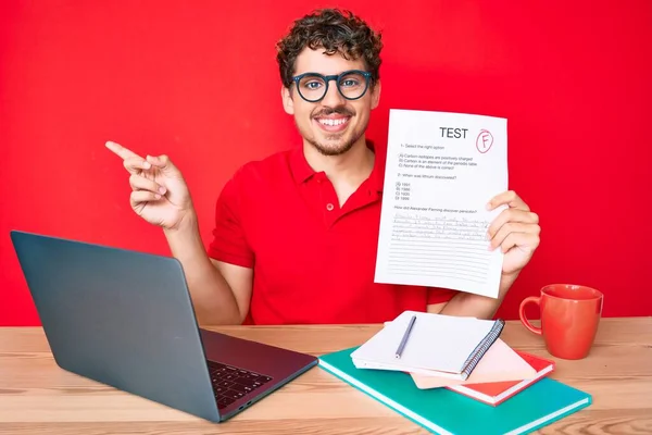 Young caucasian man with curly hair sitting on the table showing failed exam smiling happy pointing with hand and finger to the side