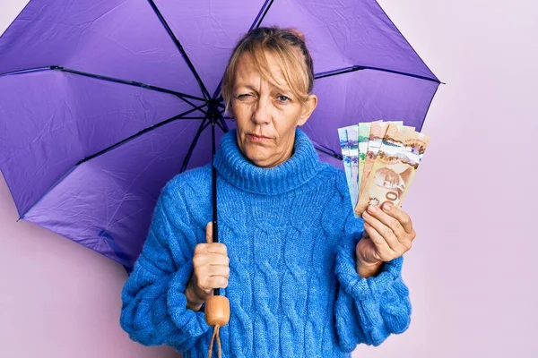 中世のブロンドの女性の傘とカナダドルの紙幣を保持懐疑的で緊張し 問題のために動揺を眉をひそめている 否定的な人 — ストック写真