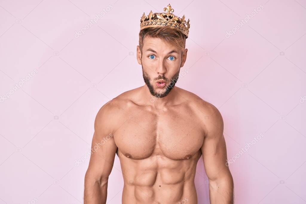 Young caucasian man shirtless wearing king crown scared and amazed with open mouth for surprise, disbelief face 