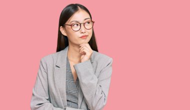 İş elbiseleri giyen genç Çinli bir kadın ciddi bir yüz ifadesi, eli çenesinde soru soran, kafa karıştırıcı bir fikir hakkında düşünceli. 