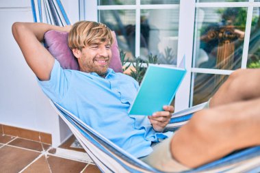 Orta yaşlı yakışıklı bir adam evinin terasında bir hamakta uzanıp kitap okuyor.