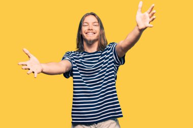 Uzun saçlı, sıradan çizgili tişört giyen yakışıklı beyaz adam sarılmak için kollarını açıp kameraya gülümsüyor. Mutluluğu kucaklayan neşeli bir ifade.. 