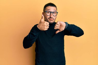 Balıkçı yaka kazak giyen, gözlük takan ve baş parmak hareketi yapan yakışıklı bir adam, anlaşmazlık ve uzlaşma ifadesi. çılgın çatışma 