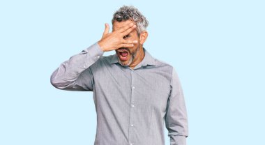 Orta yaşlı, gri saçlı, günlük kıyafetler giyen, şok içinde yüzünü ve gözlerini kapayarak utangaç ifadesiyle parmaklarının arasından bakan bir adam.. 