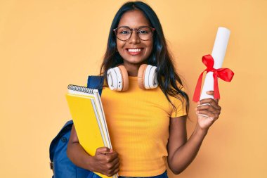 Sırt çantalı genç Hintli kız elinde diplomayla mutlu ve havalı bir gülümsemeyle gülümsüyor. diş gösterme. 