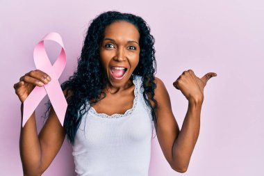 Orta yaşlı Afro-Amerikalı bir kadın pembe kanser kurdelesi tutuyor başparmağıyla yan tarafa bakıyor ve ağzı açık gülümsüyor. 