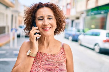 Orta yaşlı bir kadın gülümseyerek mutlu bir şekilde şehrin caddesinde yürürken akıllı telefondan konuşuyor..
