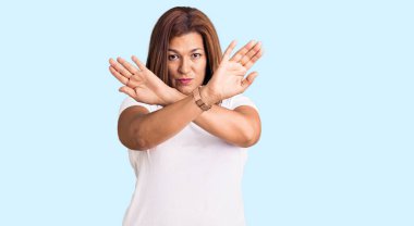 Orta yaşlı, beyaz tişört giyen Latin bir kadın. Reddedilmiş yüz ifadesi. Kollarını kavuşturuyor. 