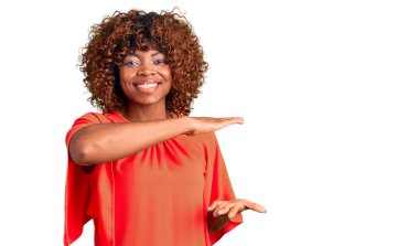 Genç Afro-Amerikalı kadın, sıradan kıyafetler giyiyor. Elleriyle büyük ve büyük bir işaret gösteriyor. Ölçü sembolü. Kameraya bakıp gülümsüyor. ölçüm kavramı. 