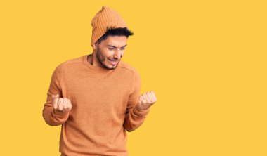 Kışlık kazak ve yün şapka giyen yakışıklı Latin Amerikalı genç adam kolları havada, gülümseyen ve başarı için bağıran, çok mutlu ve heyecanlı bir jest yapıyor. kutlama konsepti. 