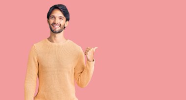 Yakışıklı İspanyol bir adam, sıradan bir süveter giyiyor, gülümsüyor, gülümsüyor ve başparmağıyla yan tarafa bakıyor.. 