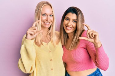 Pembe arka planda yan yana duran iki arkadaş gülümsüyor ve el hareketleriyle küçük boy işareti yapıyor ve kameraya bakıyor. ölçüm kavramı. 