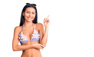 Bikini giyen, güneş gözlüklü, yüzünde kocaman bir gülümseme olan, eli ve parmağı kameraya bakan genç ve güzel bir Latin kız.. 