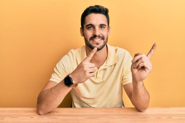 Gündelik kıyafetler giyen genç İspanyol adam masaya oturmuş gülümsüyor ve kameraya bakıyor iki eliyle ve parmaklarıyla kenarı işaret ediyor.. 