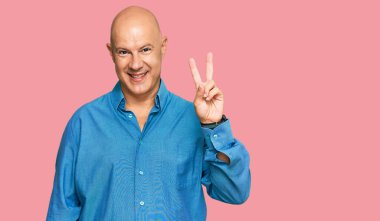 Orta yaşlı, kel bir adam, günlük kıyafetler giyip iki numaralı parmaklarıyla işaret ederken kendinden emin ve mutlu bir şekilde gülümsüyor.. 