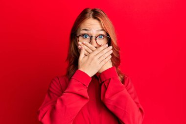 Kırmızı arka plan üzerine gündelik kıyafet ve gözlük takan genç, güzel, kızıl saçlı kadın yanlışlıkla ağızlarını kapatarak şok geçirdi. gizli kavram. 