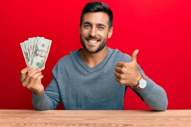 Yakışıklı İspanyol adam elinde 20 dolarlık banknotlar tutuyor mutlu ve olumlu gülümsüyor, başparmağıyla mükemmel ve onay işareti yapıyor. 