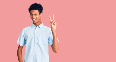 Genç Afro-Amerikalı sıradan kıyafetler giyen, gülen yüzlü, kameraya göz kırpan parmaklarıyla zafer işareti yapan genç bir adam. İki numara.. 