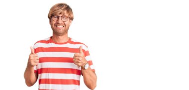 Sakallı, sıradan kıyafetler ve gözlük takan yakışıklı beyaz bir adam. Elleriyle olumlu jest yapıyor. Gülümsüyor ve mutlu. Neşeli ifade ve kazanan jest. 