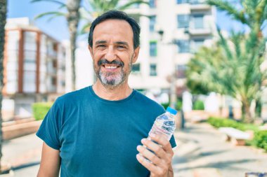 Orta yaşlı, sakallı, mutlu, açık havada su içen bir adam.