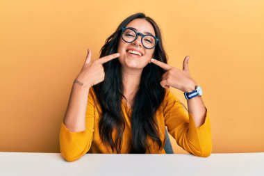 Güzel, esmer, gözlüklü ve gündelik kıyafetleri olan genç bir kadın masaya oturmuş neşeli bir şekilde gülümsüyor ve parmakları, dişleri ve ağzıyla işaret ediyor. diş sağlığı kavramı. 