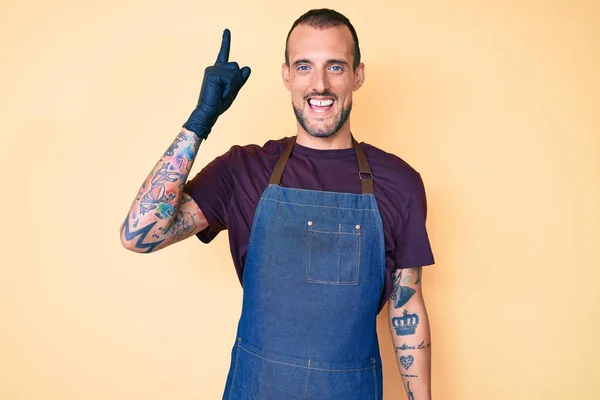 Young handsome man with tattoo wearing barber apron pointing finger up with successful idea. exited and happy. number one.