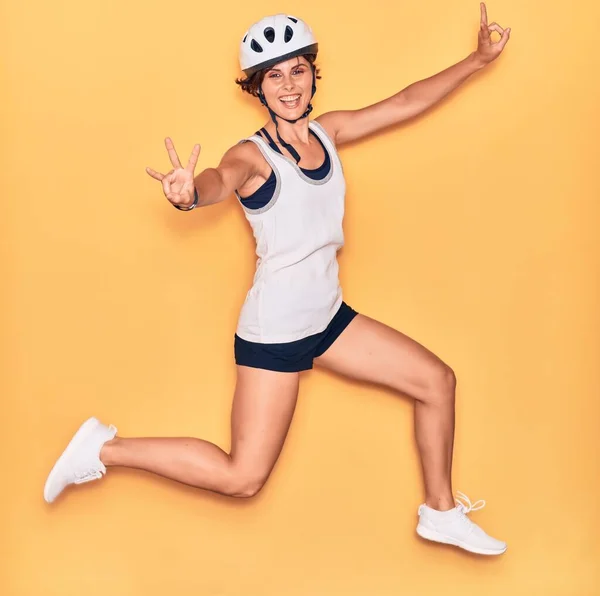若い美しいバイカーの女性が幸せな笑顔自転車ヘルメットを着用 孤立した黄色の背景の上に顔に笑顔でジャンプ — ストック写真