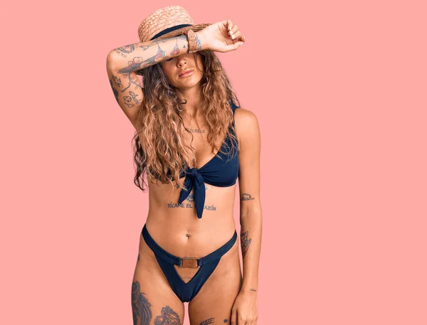 Young hispanic woman with tattoo wearing bikini and summer hat covering eyes with arm, looking serious and sad. sightless, hiding and rejection concept
