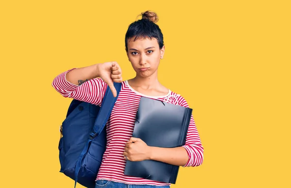 Young woman wearing student backpack holding binder with angry face, negative sign showing dislike with thumbs down, rejection concept