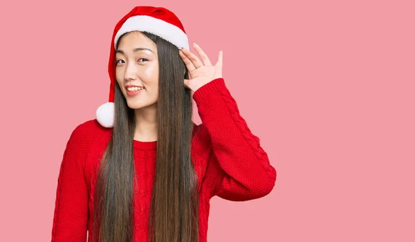 噂やゴシップに耳を傾ける耳の上に手で笑みを浮かべてクリスマスの帽子をかぶって若い中国の女性 聴覚障害の概念 — ストック写真