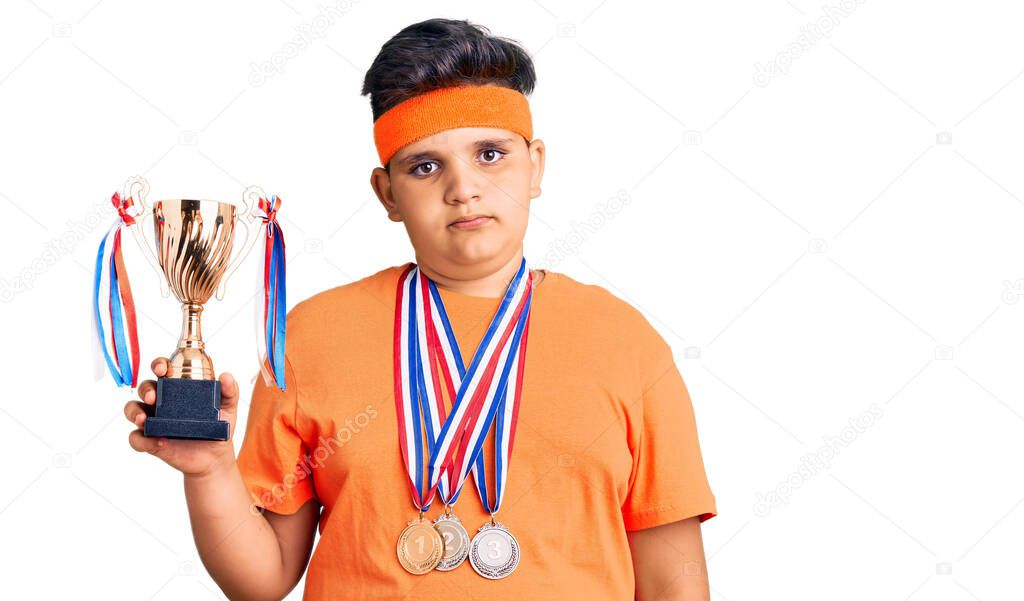 Little boy kid holding champion trophy and wearing medals thinking attitude and sober expression looking self confident 