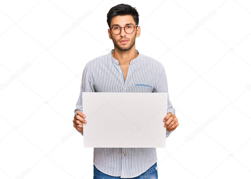 Young handsome man holding blank empty banner thinking attitude and sober expression looking self confident 