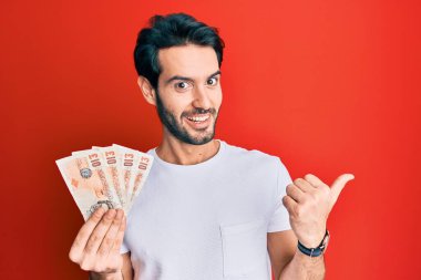 İspanyol asıllı genç adam, elinde 10 pound tutuyor. Banknotlar, başparmağıyla yan tarafa bakıyor. Ağzı açık gülümsüyor. 