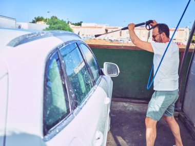 Orta yaşlı yakışıklı, günlük kıyafetler giyen ve mutlu gülümseyen güneş gözlüklü bir adam. Yüzünde gülümsemeyle ayakta durup su tabancası kullanarak araba yıkamak..