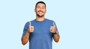 Dövmeli yakışıklı, sıradan giyinen, başarı işareti takan, elinde olumlu bir jest yapan mutlu ve gülümseyen bir adam. Neşeli ifade ve kazanan jest. 