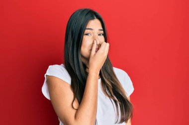 Genç esmer kadın kırmızı arka planda günlük beyaz t-shirt giyiyor pis ve iğrenç bir koku alıyor, dayanılmaz bir koku, parmakları burnunda nefesini tutuyor. Kötü kokuyor. 