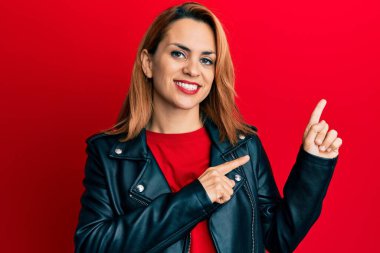 Siyah deri ceket giyen, gülümseyen ve kameraya bakan İspanyol genç bir kadın. İki eliyle ve parmaklarıyla kenarı işaret ediyor.. 