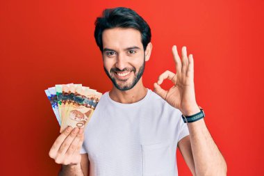 Kanada doları tutan İspanyol genç adam parmaklarıyla imza atıyor, gülümseyerek mükemmel bir sembol oluşturuyor. 