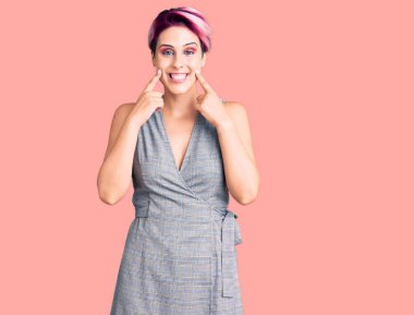 Pembe saçlı, sıradan kıyafetler giyen, ağzı açık gülümseyen, parmakları işaret eden ve zorla gülümseyen güzel bir kadın. 