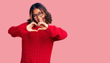 Genç ve güzel melez bir kadın kırmızı kazak ve gözlük takıyor. Sevgiyle gülümsüyor. Kalp sembolü ve şekli elleriyle gösteriyor. romantik kavram. 