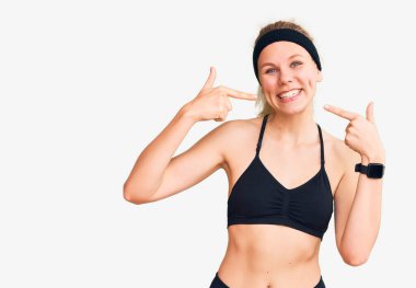 Spor kıyafetleri giyen, neşeli gülümseyen, parmaklarıyla ve ağzıyla işaret eden genç, güzel sarışın kadın. diş sağlığı kavramı. 