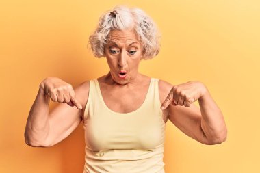 Yaşlı, gri saçlı, günlük kıyafetler giyen, parmaklarıyla işaret eden, reklam veren, şaşırmış yüzlü ve ağzı açık bir kadın. 