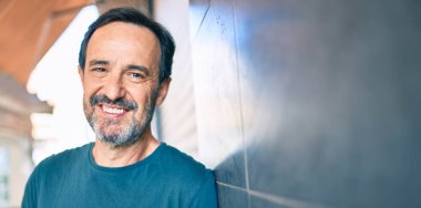 Orta yaşlı, sakallı, mutlu, açık havada duvara yaslanmış bir adam.