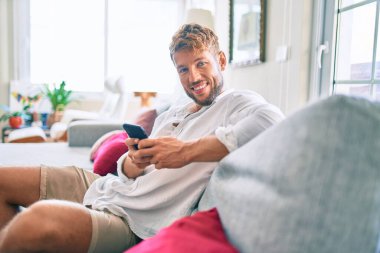 Yakışıklı beyaz adam mutlu gülümsüyor. Evdeki kanepede otururken akıllı telefon kullanıyor.
