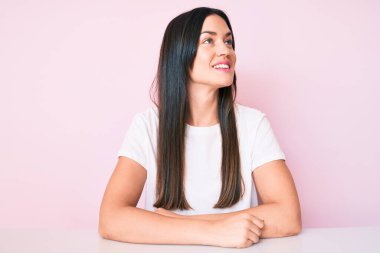 Masada oturan beyaz tişörtlü genç beyaz kadın yüzünde gülümseme, doğal bir ifade ile yan yana bakıyor. Kendinden emin gülen. 