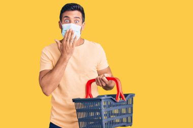 Alışveriş sepeti ve tıbbi maske takan genç İspanyol adam ağızlarını eliyle kapatıyor, şok geçiriyor ve hata yapmaktan korkuyor. şaşırmış ifade 