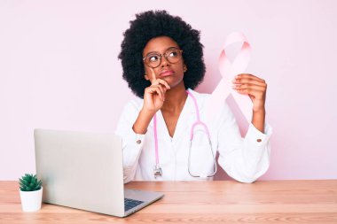 Genç Afro-Amerikan kadın doktor üniforması giyiyor pembe kanser kurdelesi takıyor ciddi bir yüz ifadesi var eli çenesinde soruyu düşünüyor, kafa karıştırıcı bir fikir. 